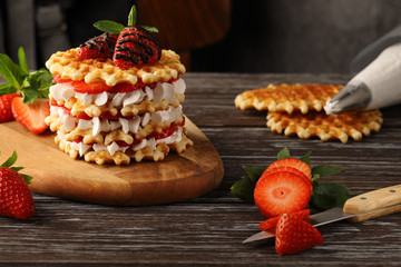 A multi-layered waffle cake with ripe strawberries and whipped cream, garnished with mint and strawberries in chocolate.