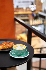 Cup of cappuccino with latte art on black metal table.With croissant.Against defocused cafe interior . Beautiful foam, greenery ceramic cups, stylish toning, place for text.