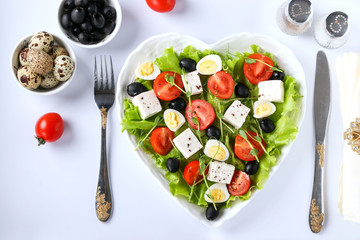 Healthy salad with cherry tomatoes, feta, quail eggs, olives and microgreens on a heart-shaped plate on a white background, healthy eating day