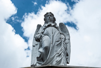 An old angel sculpture looking down in low angle shooting in top of mausoleum cemetery. Blue and cloudy sky at background