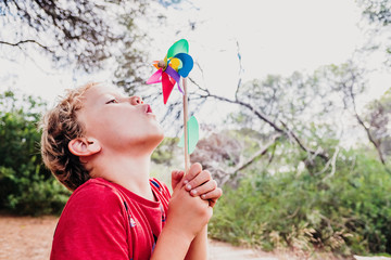 Blond boy playing in a forest with a colorful pinwheel blowing air with funny face.