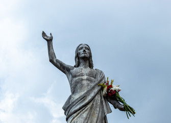 An old Jesus Christ resurrected statue with a multicolor bouquet flowers in a hand and a blue cloudy sky at background