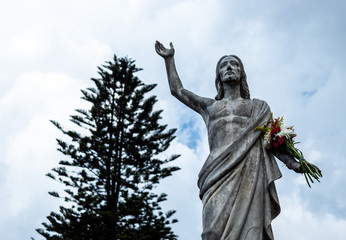 An old Jesus Christ resurrected statue with a multicolor bouquet flowers in a hand and a blue cloudy sky with a lonely tree at background
