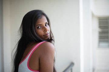 Portrait of an attractive young brunette dark skinned Indian Bengali girl in western sportswear standing on a balcony  in white urban background. Indian lifestyle