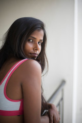 Portrait of an attractive young brunette dark skinned Indian Bengali girl in western sportswear standing on a balcony  in white urban background. Indian lifestyle