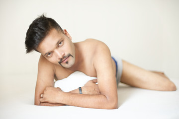 Obraz na płótnie Canvas Portrait of a confident muscular Indian brunette dark skinned macho man in bare body lying carefree on white bed and looking thoughtfully. Indian lifestyle and fashion portrait