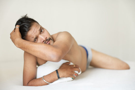 Portrait of a confident muscular Indian brunette dark skinned macho man in bare body lying carefree on white bed and looking thoughtfully. Indian lifestyle and fashion portrait