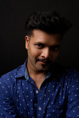 Fototapeta premium Portrait of a handsome and intelligent Indian brunette man wearing blue shirt with white stars sitting on a chair. Indian lifestyle and fashion portrait
