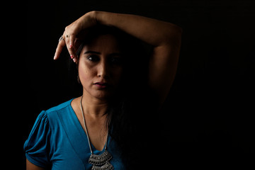 Obraz premium Close up face portrait of a beautiful Indian Bengali brunette woman in light and shadow before a black copy space background wearing a blue top. Indian lifestyle and fashion portrait