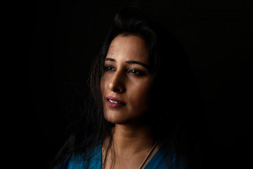 Fototapeta premium Close up face portrait of a beautiful Indian Bengali brunette woman in light and shadow before a black copy space background wearing a blue top. Indian lifestyle and fashion portrait