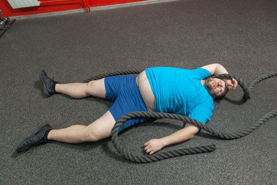 Overweight man is lying on the floor exhausted after performing battle rope exercise in the fitness gym