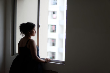 Backside portrait of a beautiful Indian Bengali brunette woman wearing a black western dress looking thoughtfully while standing near a window. Indian lifestyle and low key fashion portrait