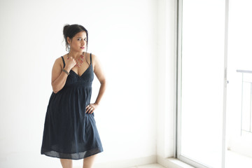 Portrait of a beautiful Indian Bengali brunette woman wearing a black western dress looking thoughtfully while standing near a balcony with a glass door. Indian lifestyle and fashion portrait