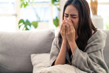 Sick Asian young woman sneeze at home on the sofa with a cold, she is covering with a blanket and blowing her nose. medical and health concept. Used tissue paper, medicine and water are put on table.