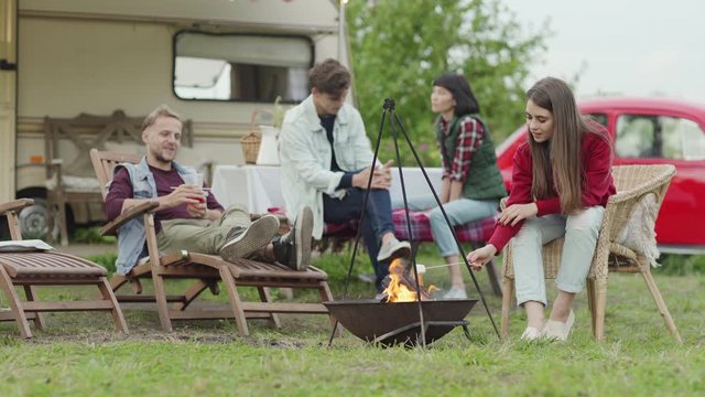 Young woman in casual outfit roasting marshmallow over fire and speaking with man while sitting on chair near chatting multiracial friends in countryside