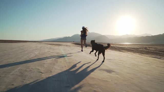 Fit female runs with her funny dog in dry desert at sunrise rapid slow motion