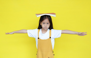 Unhappy asian child girl with book on head isolated on yellow background. Portrait of punished...