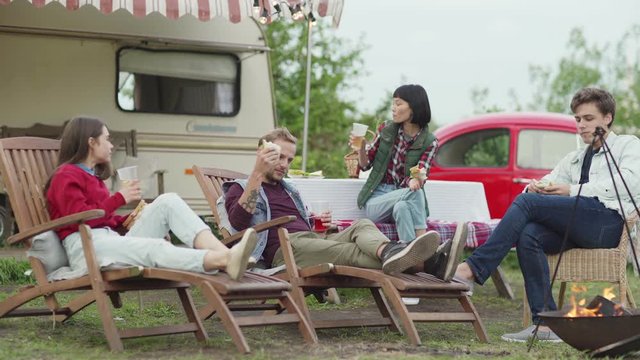 Group of diverse young people eating sandwiches and drinking alcohol while resting and chatting during picnic near caravan and car in nature
