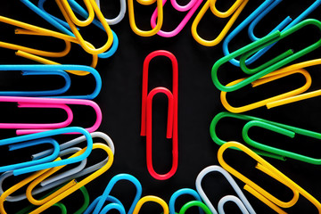 Red paper clip surrounded by colorful paper clips on black background.
