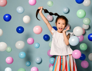 Fototapeta na wymiar Cheerful frolic asian kid girl is having fun surrounded by colorful air balloons holding up her long black plait