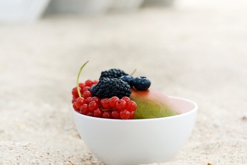 Summer fruits in a bowl on a background of sea sand. Summer vacation on the beach. Ripe whole  mango and berries of blackberry, raspberry, red currant in a bowl. Breakfast in the sand