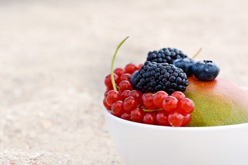 Summer fruits in a bowl on a background of sea sand. Summer vacation on the beach. Ripe whole  mango and berries of blackberry, raspberry, red currant in a bowl. Breakfast in the sand