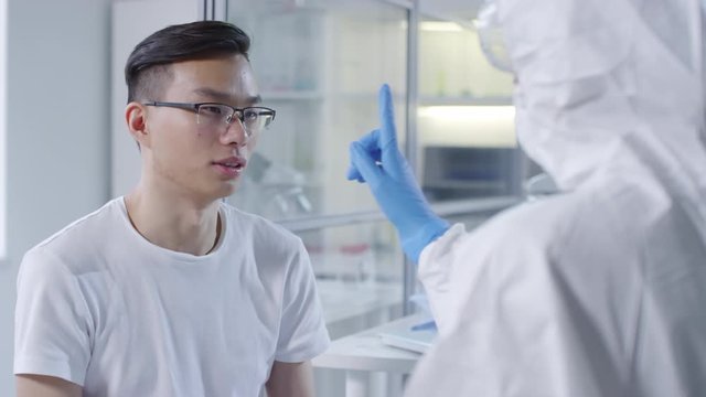Medium shot of unrecognizable doctor in disposable coveralls, safety goggles and gloves and doing neurological examination of infected male Asian patient in glasses