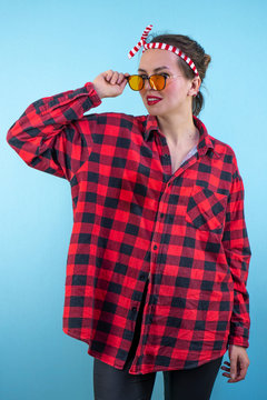 Beautiful young woman with pin-up makeup, wearing fashionable glasses, posing in studio. Isolate on a blue background. Girl in a checkered shirt and orange stylish glasses.