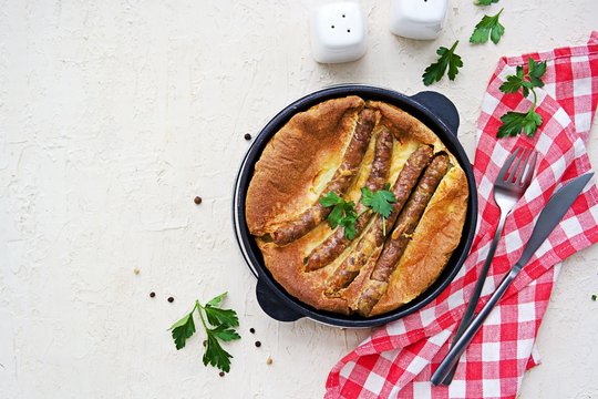 Pork sausages baked in pastry for Yorkshire pudding in a serving cast-iron skillet on a light concrete background. Toad in a hole recipe. British cuisine. Top view, copyspace