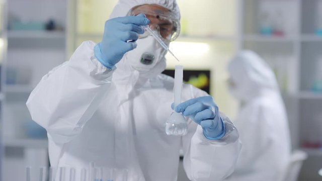 Tilt up shot male chemist in protective coveralls, respirator mask, goggles and gloves using pipette and dropping liquid solution into flask, then looking at chemical reaction