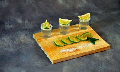 A wooden stand with three glasses of tequila with salt and lime slices stands on a gray abstract...