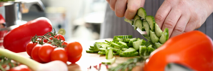 Male cook in apron cut green cucumber on wood board concept aganist kitchen background