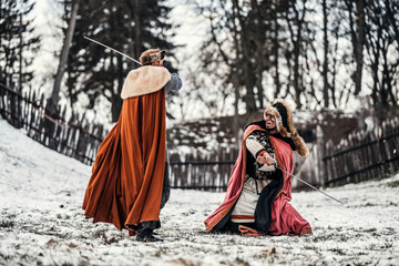 Battle of two knights in colored robes and hats near forest and wooden fortress. Knights fight in...