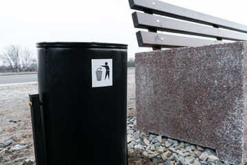 Trash can stands on the street near the bench. Trash bin close-up.