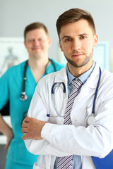 Fototapeta premium Portrait of handsome man in white uniform with stethoscope. Smiling doc posing at workplace in clinic office. Medical treatment concept. Blurred background