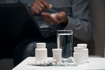 Overworked businessman sitting on the sofa and taking medication. Pills and glass of water near him.