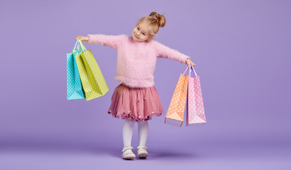 Sale of childrens products. Portrait of fun little child girl  on purple background holding shopping bags, package. Looking at the camera. Banner. Space for text.