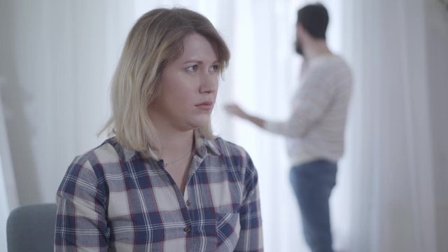 Young pretty Caucasian wife listening carefully to husband talking on the phone at the background. Portrait of jealous woman spying after spouse indoors. Relationship, marriage, trust, reliance.