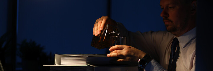 Portrait of tired businessman sitting indoors and pouring alcohol in glass. Man relaxing after intense workday with pile of paper folders full of corporation documents. Night at office concept