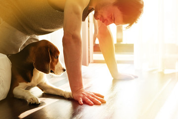 Smiling bearded man doing exercises push ups against the window at home with his dog