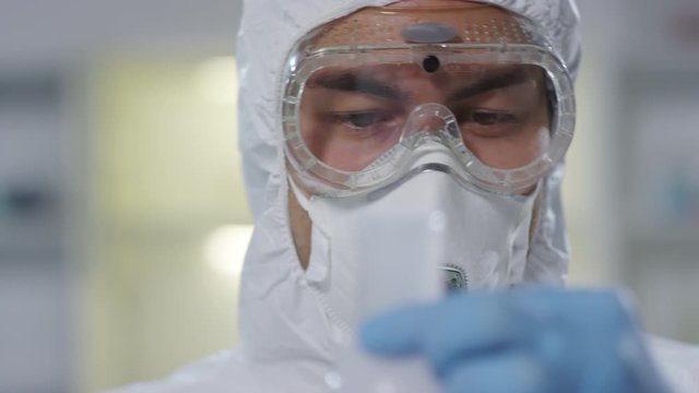 Close up of male scientist in disposable coveralls, goggles, respirator mask and gloves holding flask and observing chemical reaction