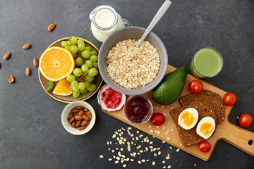 food, eating and breakfast concept - oatmeal cereals in bowl, fruits, almond nuts, glass of juice and jug with milk on slate stone table