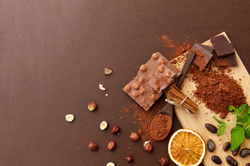 sweets, confectionery and culinary concept - chocolate with hazelnuts, cocoa beans, and powder with dry orange and cinnamon on wooden board with spoon on brown background