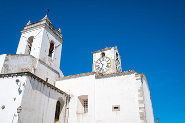 Fototapeta premium Bright morning view of traditional whitewashed Igreja de Santa Maria do Castelo church with clock tower under blue Mediterranean sky in Faro, Algarve, Portugal