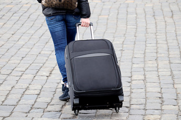 Woman in jeans walking with a suitcase on wheels. Female legs and luggage on the cobbled street,...