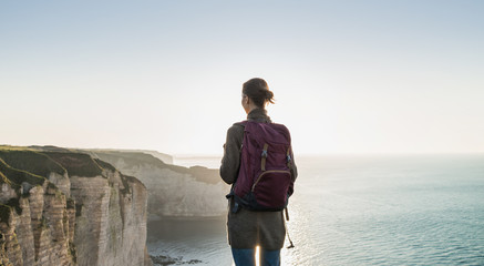 Young woman traveler with backpack looking at sea in Normandy, France over beautiful cliffs...