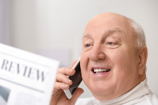 Portrait of elderly man talking by phone while reading newspaper at home