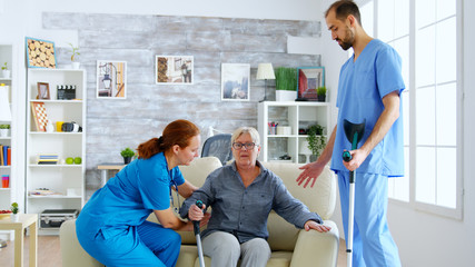 Female doctor and her assistant helping old woman with crutches to stand up from the couch