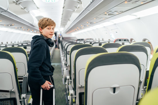 Smiling blonde hair teenager portrait staying in aircraft corridor with headphones with cabin trolley bag. Kids traveling or unaccompanied child in aircraft concept image.