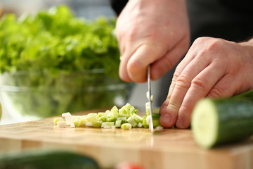 Cook holds knife in hand and cuts on cutting board green onions for salad or fresh vegetable soup with vitamins. Raw food and vegetarian recipe book in modern society popular concept.